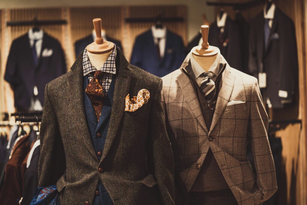 Two mannequin displays showcasing stylish men's suits in a luxurious store setting. The left mannequin features a dark herringbone tweed suit with a blue waistcoat, patterned orange tie, and matching pocket square. The right mannequin is dressed in a beige checkered suit with a gray sweater, striped tie, and white pocket square, surrounded by other elegant clothing in the background.